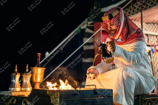 A man sits in front of an open fire at night, wearing traditional Saudi attire, including a thobe, ghutra, and agal. He is pouring Arabic coffee from a dallah into a small cup. In the background, there is a brightly colored Arabian tent, and there are additional coffee pots near the fire.