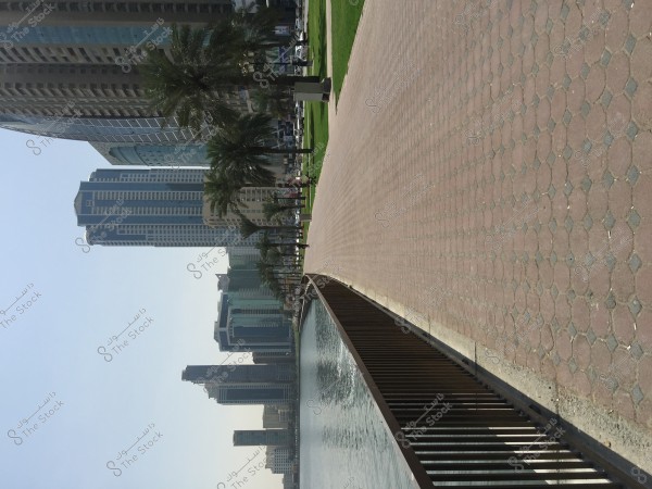 A paved brick walkway along a waterfront, with tall buildings in the background and palm trees lining one side of the path. The sky is clear and the weather is sunny, adding a serene feel to the urban scene. Cars can be seen parked along the road in the distance.