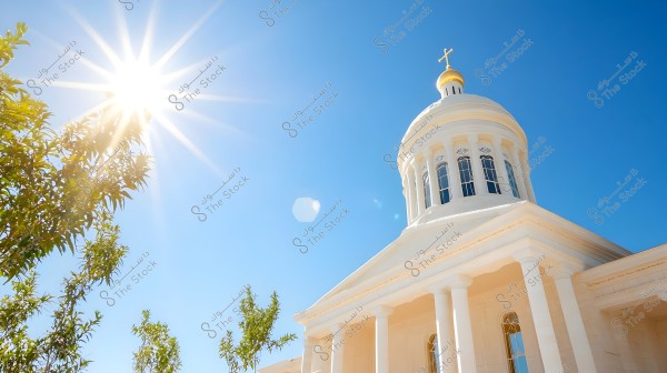 Image showing a white church dome capped with a golden cross, set against a sunny blue sky. The bright sunlight shines from the upper left, reflecting on tree leaves in the left foreground of the photo.
