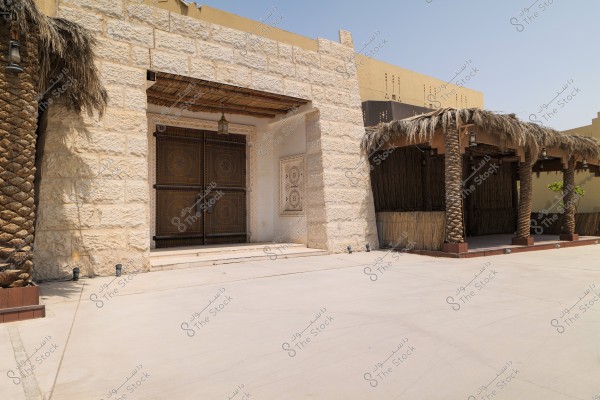 The image shows a traditional building made of white stone, featuring a large ornate wooden gate. On the side of the building, there is a colonnade made from palm tree pillars and a palm frond roof, giving it a traditional Arabic feel. The background shows a clear blue sky.