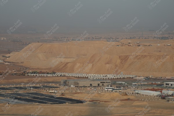 A wide view of a large construction site in a desert area. Vast expanses of sandy soil are visible with construction equipment such as excavators and trucks spread across the site. Dirt mounds and a backdrop of distant desert haze are present, with several temporary buildings in the foreground.