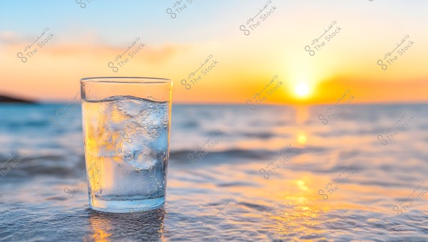 An image of a clear glass of water with ice cubes, placed on a beach. The background features a golden sunset reflecting on the surface of the water, creating a picturesque scene.