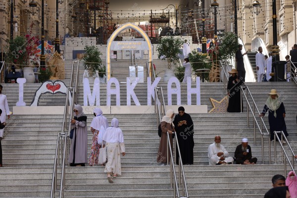 The image shows an outdoor staircase adorned with large letters spelling \"I ❤️ MAKKAH\" in the city of Mecca. People are seated and walking, wearing various attire including traditional robes and head coverings. In the background, building entrances with illuminated arches and hanging decorative lights are visible. Flowers and plants adorn the sides, creating a lively atmosphere filled with visitors coming and going.