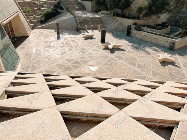 An image of an open area covered with interlocking panels creating a geometric pattern. The location is surrounded by stairs leading upwards and corners with small trees and plants. There are star-shaped concrete benches in the courtyard. The place looks modern and designed in a contemporary style.