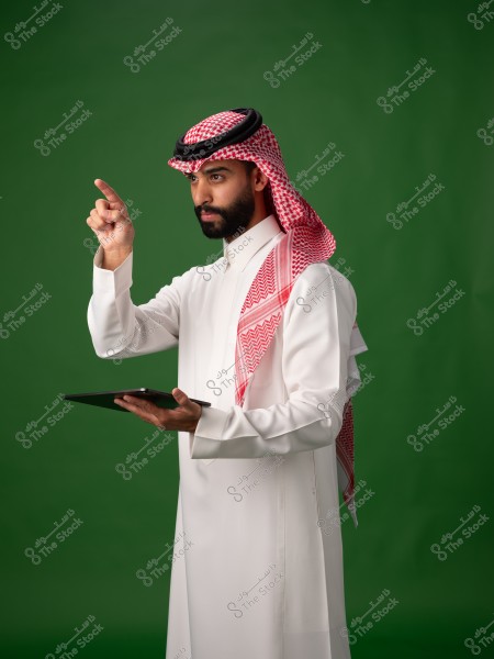 Image of a man wearing a traditional Saudi white thobe with a red and white checkered ghutra and black agal, standing against a green background. He is holding a tablet in his left hand and pointing with his right index finger, focusing on something ahead.