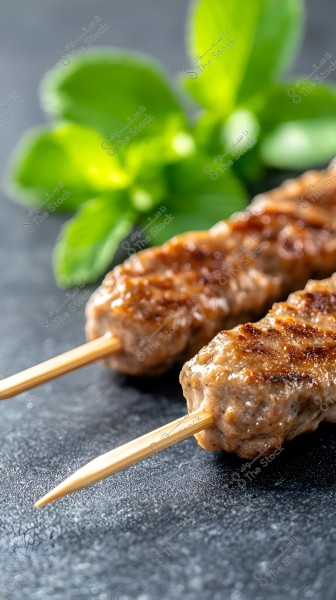 Two skewers of grilled meat shaped like kebabs placed on a dark surface. In the background, there are bright green mint leaves, adding a touch of color and freshness.