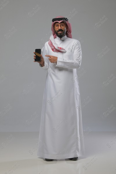 صورة لرجل يرتدي الثوب السعودي التقليدي الأبيض والشماغ الأحمر مع العقال الأسود. يقف ويشير بيده إلى هاتف ذكي يحمله في اليد الأخرى. يظهر في الخلفية لون رمادي محايد مما يبرز تفاصيل الزي والهاتف بشكل واضح.