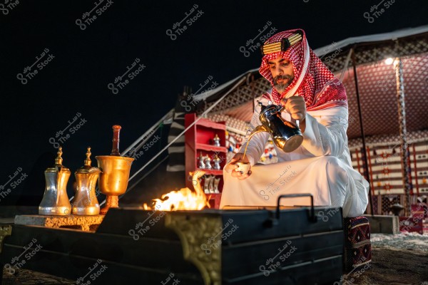 رجل يرتدي الزي التقليدي العربي, جالسًا أمام خيمة عربية مزخرفة, يقوم بصب القهوة العربية من دلة معدنية إلى فنجان صغير. يوجد أمامه نار مشتعلة وتُرى أواني معدنية أخرى على طاولة قريبة.