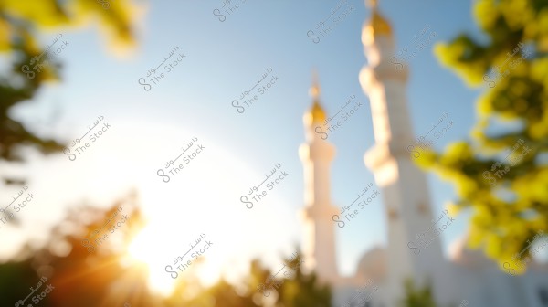 A blurred image of a mosque with two white minarets, with bright sunlight glowing between green trees. The sky is clear blue, and the scene appears serene.