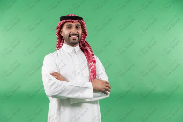 رجل سعودي  مرتديًا الثوب الأبيض وشماغ أحمر، يقف بابتسامة وأذرعه متقاطعة على خلفية خضراء.