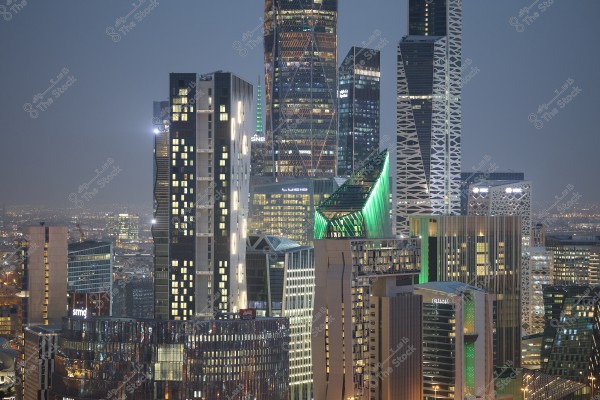 A nighttime image of Riyadh\'s skyline in Saudi Arabia, featuring a collection of modern skyscrapers illuminated with vibrant lights. The tall, architecturally diverse buildings dominate the scene, lighting up the surrounding cityscape. There is a contrast between different structural designs and vivid lighting, conveying a sense of development and vibrancy.