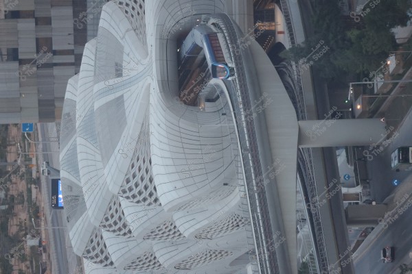 A modern and striking building with an innovative design that combines traditional Arabic architecture with modernity. The exterior features intricate lattice patterns, with a high-speed train running over a raised bridge above. Some trees and cars can be seen on the street below.