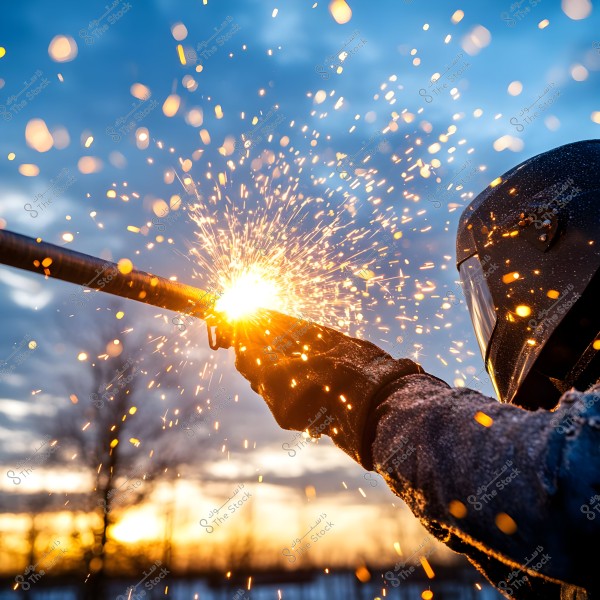 An image of a welder wearing a black helmet and protective goggles, working outdoors. Bright sparks fly from the welding machine as the welder works during sunset. Trees are visible in the background against an orange and blue cloudy sky.