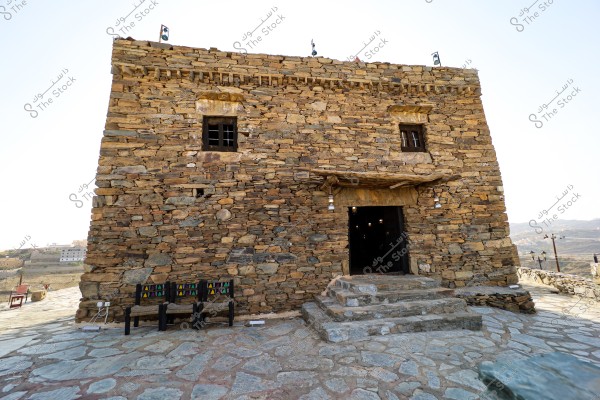 Image of a historical stone building in a mountainous area, possibly an old fortress or house. The building is constructed with large stones and features small metal windows. In front of the main entrance, there is a small stone staircase and hanging lanterns. On the left side, there are traditional wooden chairs and a small table. The surrounding environment indicates a high-altitude area with views of distant buildings and mountainous terrain.