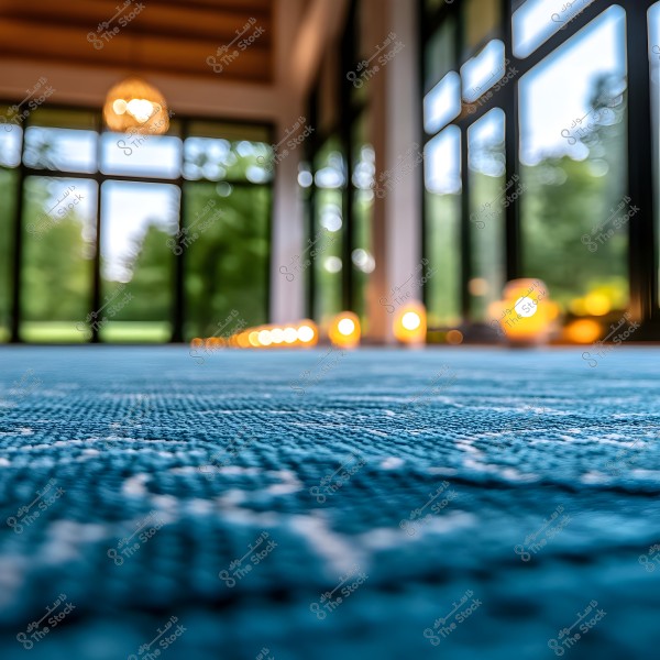 An image showing an interior view of a room with a blue patterned carpet on the floor. In the background, a series of glowing lights are visible on the floor, with large glass windows providing a view of trees outside.