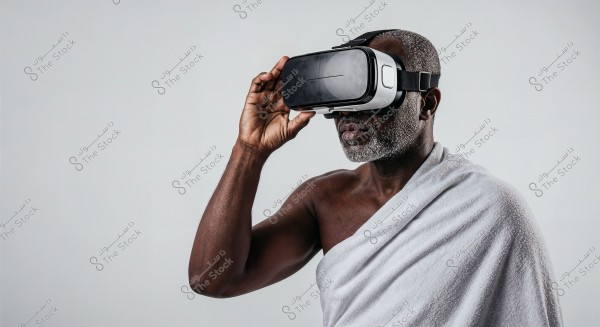 Image of an elderly man wearing a virtual reality headset, with his body draped in a white piece of cloth resembling traditional attire. The background is a soft gray, highlighting the details of the person and the VR headset.