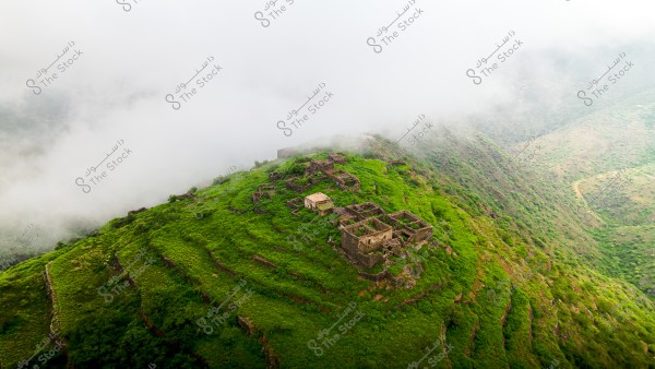 An aerial view of green hills covered with dense grasses and surrounded by thick fog. In the center of the image, remnants of ancient stone structures are scattered across different levels of the land. The scene conveys a sense of tranquility and antiquity.