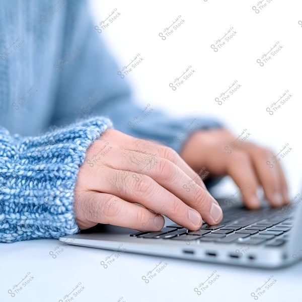 Image of a pair of hands on a laptop keyboard. The hands are wearing a knitted blue sweater. The focus is on the details of the hands and the knitting, with the device placed on a light-colored flat surface.