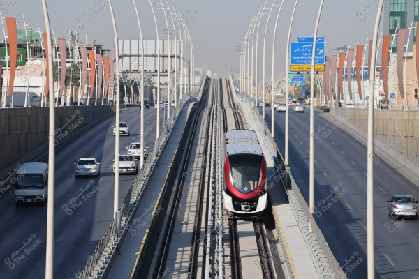 The image shows a modern train traveling on an elevated railway track between two busy highways in a large city. Tall street lights line the roads, and several cars are driving alongside the train. Road signs in Arabic and English indicate different directions and destinations. Modern buildings and palm trees can be seen in the background.