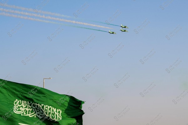 Three airplanes flying in formation in the blue sky, trailing colored smoke. The green Saudi flag is visible in the lower corner of the image.
