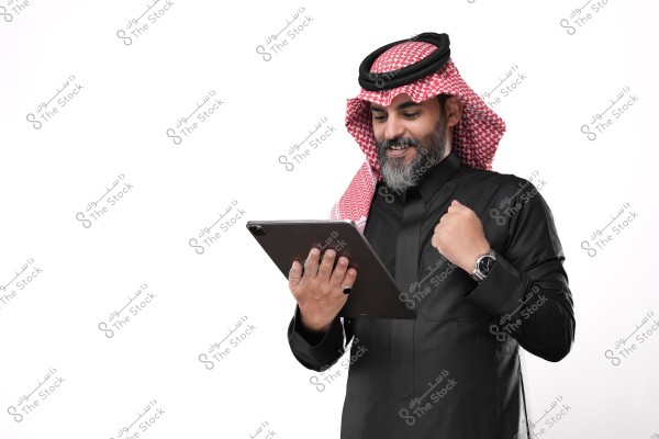 رجل عربي يرتدي ثوبًا تقليديًا وغطاء رأس, ينظر إلى لوحة إلكترونية مبتسمًا وخلفة خلفية بيضاء.