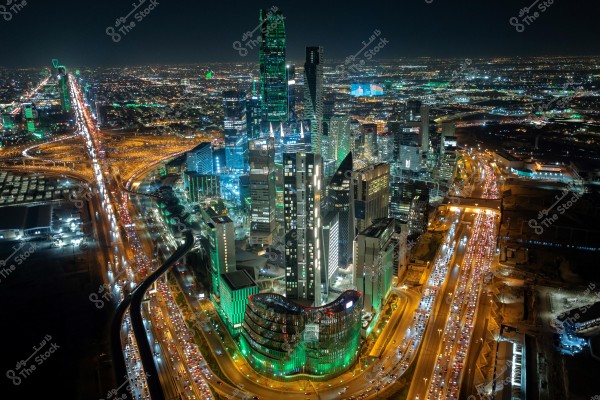 An aerial view of Riyadh at night, showcasing high-rise towers and buildings illuminated with bright lights. The crowded streets with traffic stretch across the city, with recognizable landmarks such as the Kingdom Tower visible in the background. Some buildings are lit with green lights, adding a vibrant ambiance to the city.