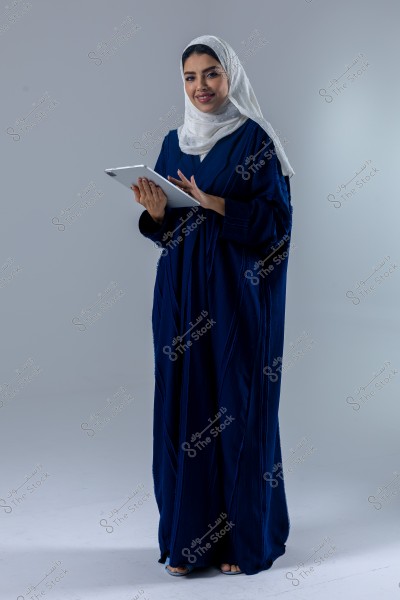 Image of a woman wearing a dark abaya and a white headscarf, holding a tablet in her hands. She stands against a light grey background and appears to be smiling. The clothing suggests a Middle Eastern style, possibly from Saudi Arabia.