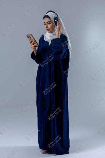 An image of a woman wearing a blue abaya and a white hijab. She is holding a phone in her hand and wearing headphones. The background is neutral, and she is focused on the phone.