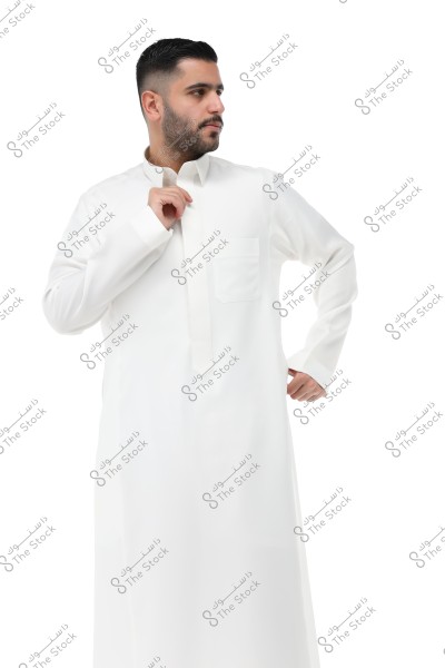 A portrait of a man wearing a traditional white thobe. The thobe features a long collar and long sleeves. The background is white, highlighting his attire. The man is looking to the left and holding the collar of the thobe with his hand.