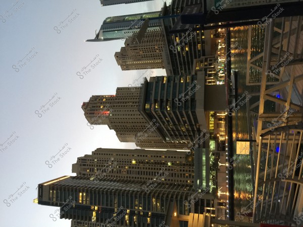 An image of a cluster of high-rise towers in a modern urban area, with buildings lit up at night and light reflecting on the adjacent waterway. The backdrop includes a clear sky, and the area seems bustling with activities along the waterfront promenade.