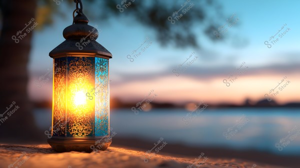 A traditional lantern illuminated in the evening with intricate blue and gold stained glass. The lantern is placed on a flat surface, with the background featuring a natural scene of a lake or river under a sunset sky with warm hues of blue and orange.