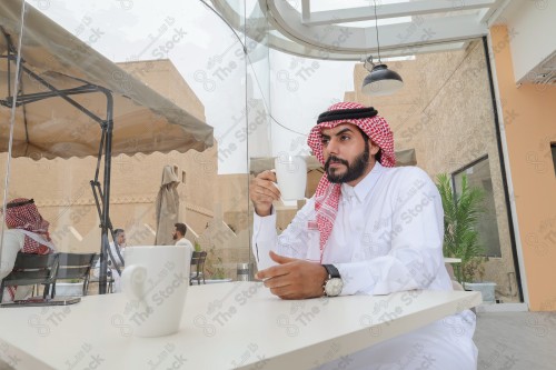 رجل سعودي يرتدي الزي السعودي التقليدي يضع ساعة على معصمه بينما يحتسي كوب من القهوة أثناء النهار