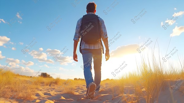 Image of a person walking in the desert during sunset. They are wearing a plaid shirt and jeans, carrying a black backpack. The sky is clear with some scattered clouds, surrounded by wild grass and sand.