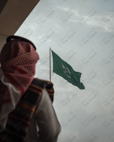 A Saudi man in traditional Saudi dress stands tall in front of the Saudi flag above a building in Rijal Almaa village during the day in the Asir region. A Saudi man celebrates the National Day, the Saudi National Day.