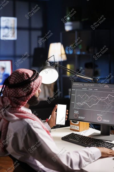 رجل يرتدي الزي التقليدي الخليجي يجلس أمام مكتب في مكتب حديث. يرتدي شماغاً أبيض وأحمر مع عقال أسود, ينظر إلى شاشة كمبيوتر تعرض تقريراً سنوياً يحتوي على مخططات ورسوم بيانية. يمسك بيده هاتفاً ذكياً بشاشة بيضاء. الأجواء هادئة حيث يبدو المكتب بإضاءة دافئة وديكور عصري في الخلفية.