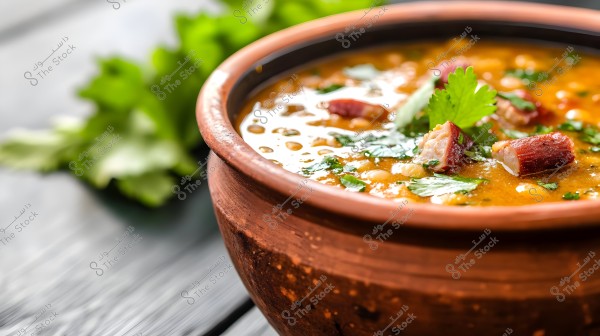 A clay pot containing a soup made of lentils and vegetables with small cubes of meat, garnished with fresh cilantro leaves. In the background, there is a blurred view of green plants on a wooden table.