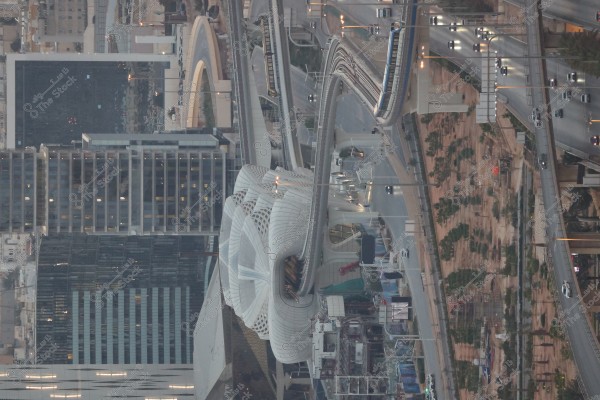 The image displays an urban scene featuring a modern, architecturally complex building resembling a train station or public transport hub. The building has a unique white geometric design with curved surfaces, encompassed by pedestrian bridges and roads busy with cars. Skyscrapers and tall buildings form the background. The photo appears to be taken at dusk, with some lights illuminated.