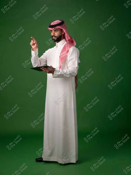 صورة لرجل يقف أمام خلفية خضراء, يرتدي ثوبًا أبيض وغطاء رأس تقليدي أحمر وأبيض يُعرف بالغترة والعقال. يبدو أنه يشير بيده اليمنى بينما يمسك بجهاز لوحي بيده اليسرى.