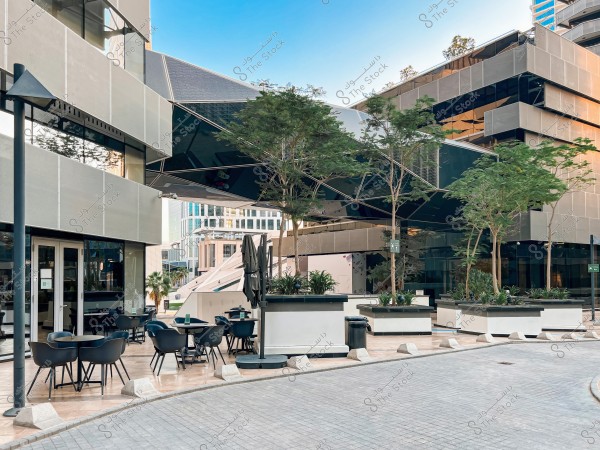 Image of a modern building with a contemporary design, featuring large windows and glass walls reflecting the blue sky. In the foreground, there is an outdoor seating area with black tables and chairs, surrounded by trees planted in white planters. Other tall buildings can be seen in the background.