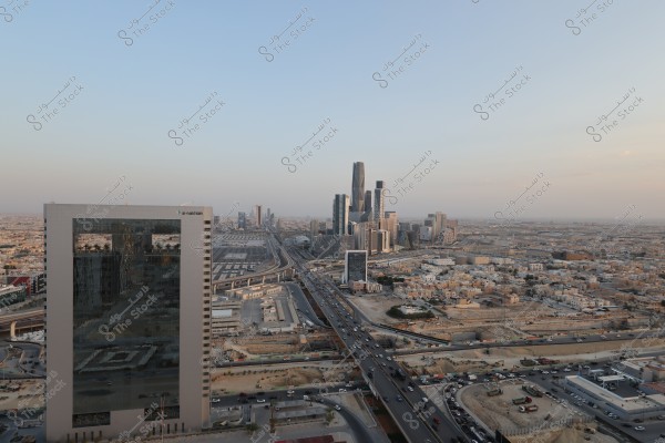 Aerial view of Riyadh city showing tall buildings and modern skyscrapers along the main road. There is heavy traffic and residential buildings visible in the horizon under a clear blue sky.