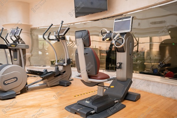 A gym featuring various exercise equipment. In the foreground, there is an arm workout machine with a display screen, while cardio machines such as stationary bikes are visible in the background. The floor is wooden and there is a mirror on the back wall reflecting the equipment.