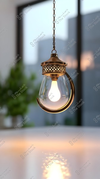 A hanging lamp with a crescent design made from golden metal and clear glass. The lamp is brightly lit, reflecting its light on a shiny surface. In the background, there are some indoor plants and soft silhouettes of buildings or glass windows.