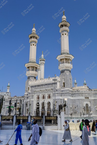 The image shows the Grand Mosque in Mecca, Saudi Arabia, featuring prominent white minarets and intricate Islamic architecture. The background has a clear blue sky, and several people are visible wearing traditional clothing, performing the Tawaf around the Kaaba.