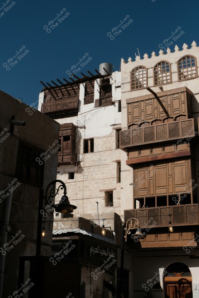 The image shows traditional buildings with old Arabic architectural style, featuring ornate wooden windows in brown. There is a sign that reads \"عطارة المغرب العربي\" and hanging street lamps that add a warm atmosphere to the scene. The sky is clear blue, reflecting a sunny weather.