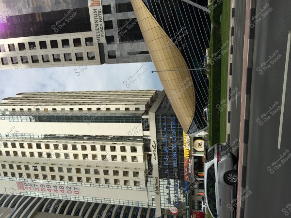 The image shows a high-rise building located beside a dual carriageway in an urban area. The building is named \"Millennium Plaza Hotel\" and features several floors with glass windows. Beside it, there is a distinctive architectural structure resembling a dome made of glass and metal. At the bottom of the buildings, there is signage for a medical center and various shops. A gray car is parked nearby.