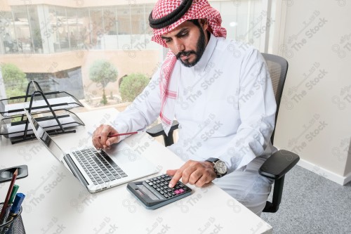 رجل سعودي يرتدي الزي السعودي التقليدي يعمل في مكتب بواجهة زجاجية ويستخدم قلم رصاص لتدوين الملاحظات