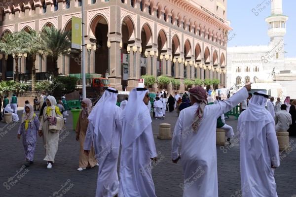 The image shows a group of people walking around an outdoor square next to a large building adorned with arches and columns. The men in the foreground are wearing traditional Gulf attire, including white thobes and headscarves. The women are in long dresses and head coverings. Palm trees and traditional Islamic architecture are visible in the background. The image appears to be taken in a public area in Saudi Arabia.