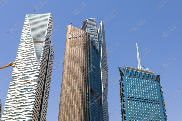 صورة لأربعة أبراج ناطحات سحاب في مدينة الرياض, السعودية. الأبراج تتميز بتصميمات معمارية حديثة ومختلفة. يظهر البرج البني الطويل في الوسط باسم \"بنك الرياض\", بينما البرج الأزرق على اليمين يضم شعار \"SNB\". السماء صافية وزرقاء في الخلفية.