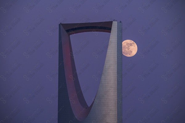 An aerial shot of the Kingdom Tower with the moon behind it in the city of Riyadh at night. The sky is clear, towers, skyscrapers, landmarks, streets and roads.