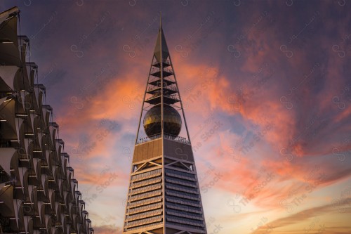 An aesthetic shot showing Al-Faisaliah Tower, one of the buildings and landmarks of Riyadh city and next to it is the King Fahd Library. The sky appears orange at sunset.